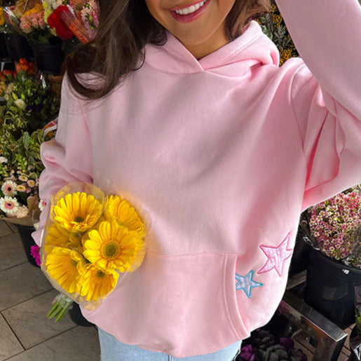 Donna con felpa rosa e fiori gialli, circondata da piante e fiori in un negozio. Felpa con stelle ricamate, atmosfera primaverile e vivace.