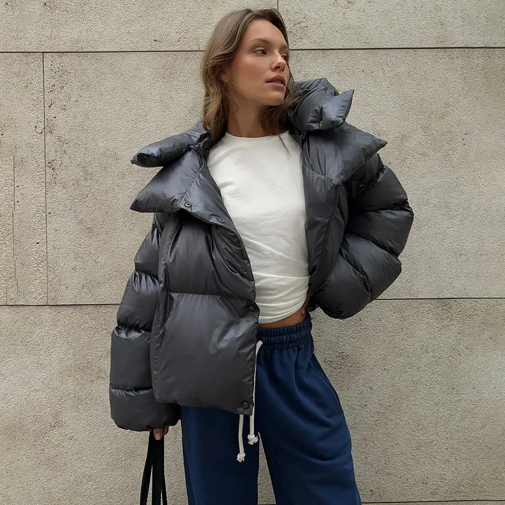 Donna con giacca piumino grigia oversize, maglia bianca e pantaloni sportivi blu, posa contro un muro. Moda invernale casual e trendy.