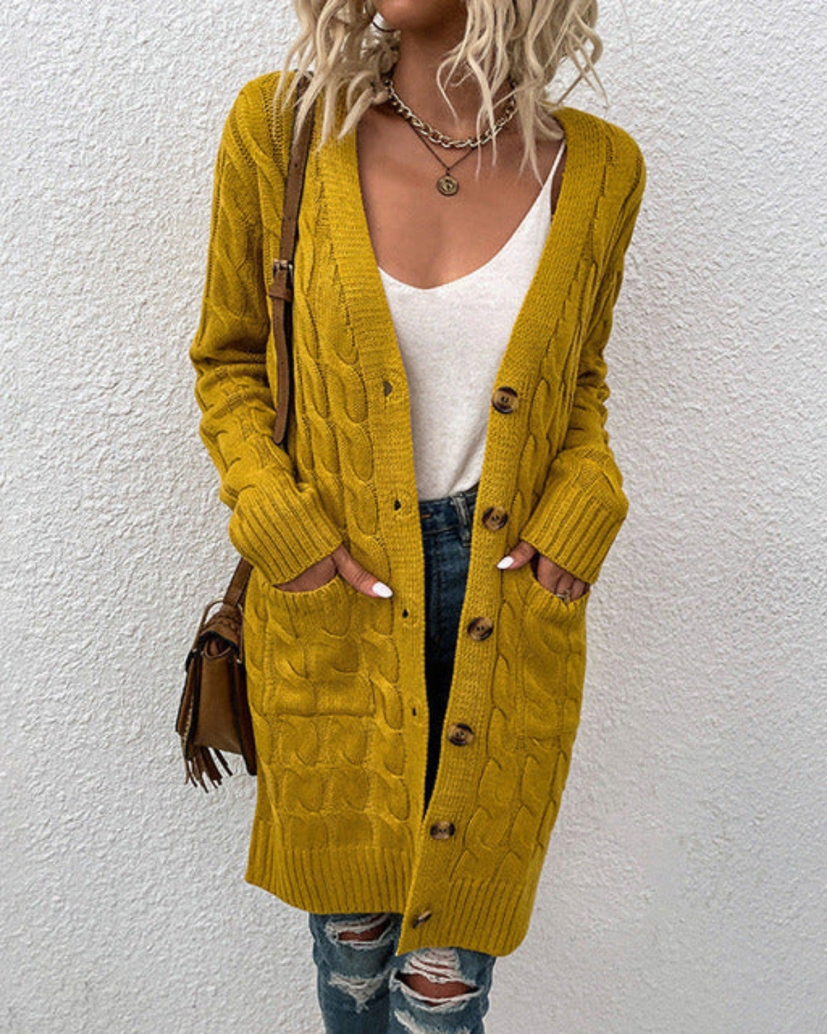Donna con cardigan giallo senape a maglia, bottoni in legno, indossa jeans strappati e top bianco, borsa marrone a tracolla, collana dorata.