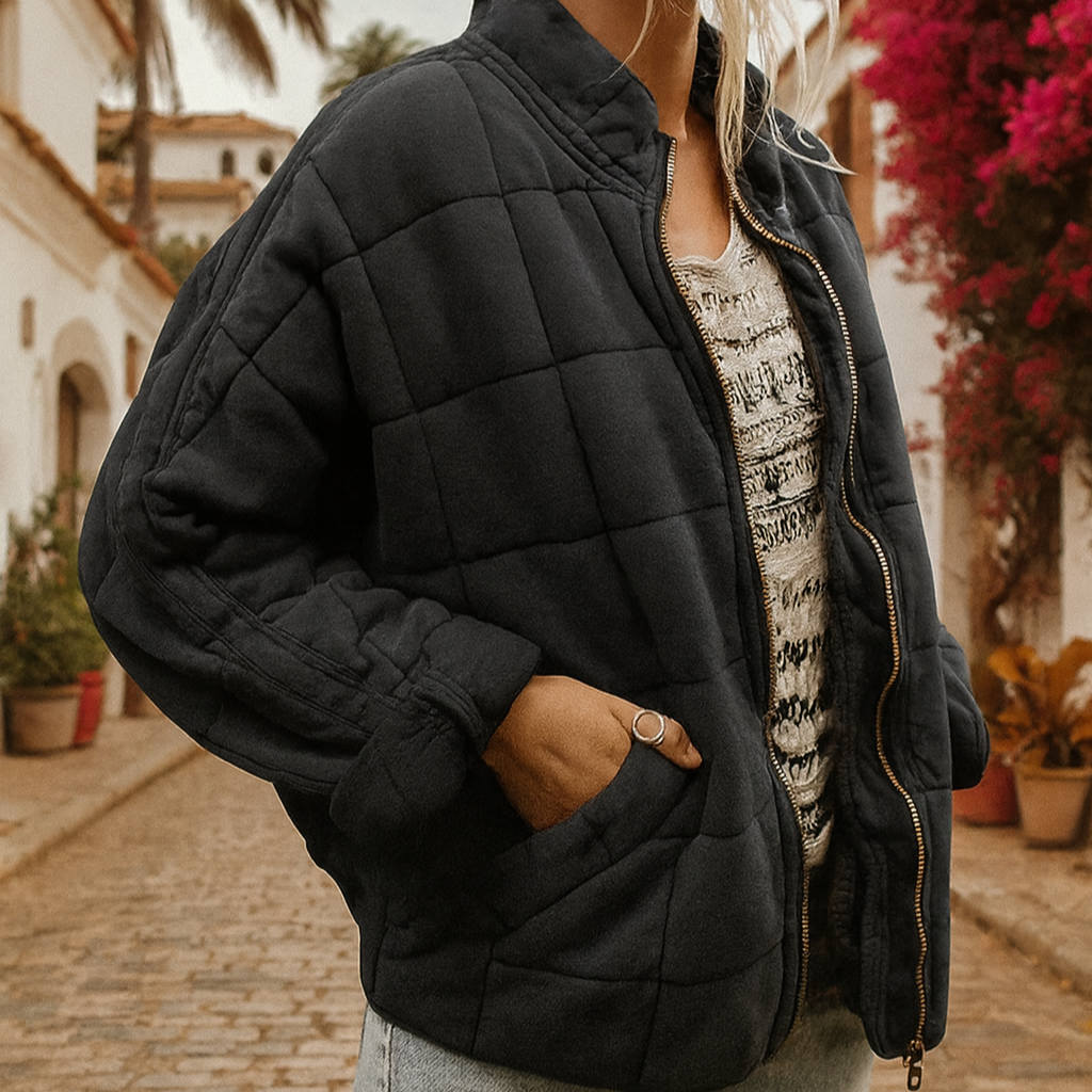 Donna con giacca trapuntata nera su strada acciottolata, sfondo di edifici bianchi e fiori colorati. Moda autunnale, stile casual urbano.