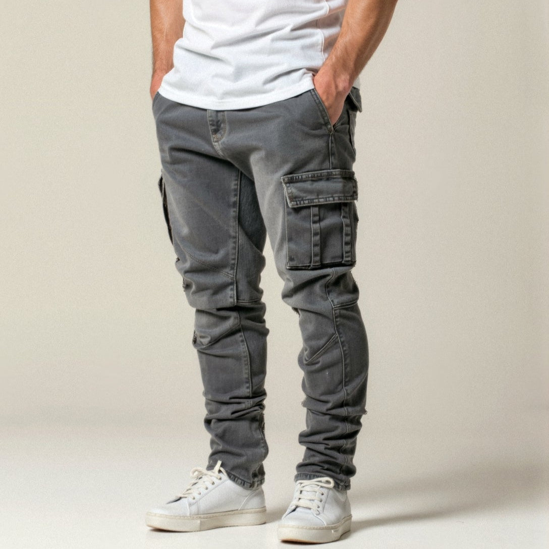 Pantaloni cargo grigi da uomo, tasche laterali, abbinati a scarpe da ginnastica bianche e maglietta bianca. Stile casual moderno.