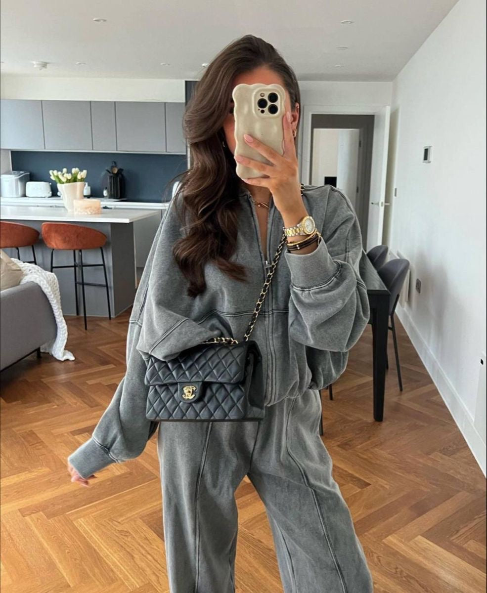Donna in tuta grigia, borsa a tracolla nera, scatta selfie in salotto moderno con pavimento in legno e cucina sullo sfondo.
