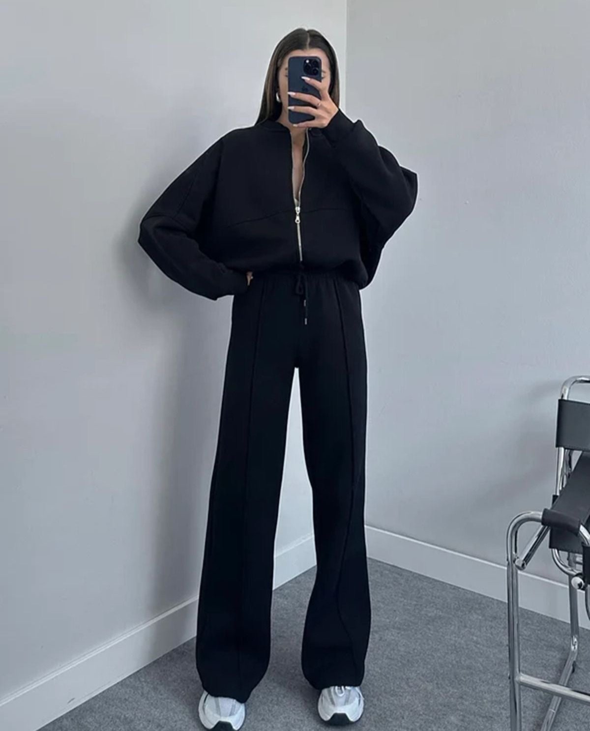 Donna in tuta nera con zip, pantaloni larghi e scarpe bianche, scatta un selfie in una stanza minimalista. Moda casual, abbigliamento sportivo.