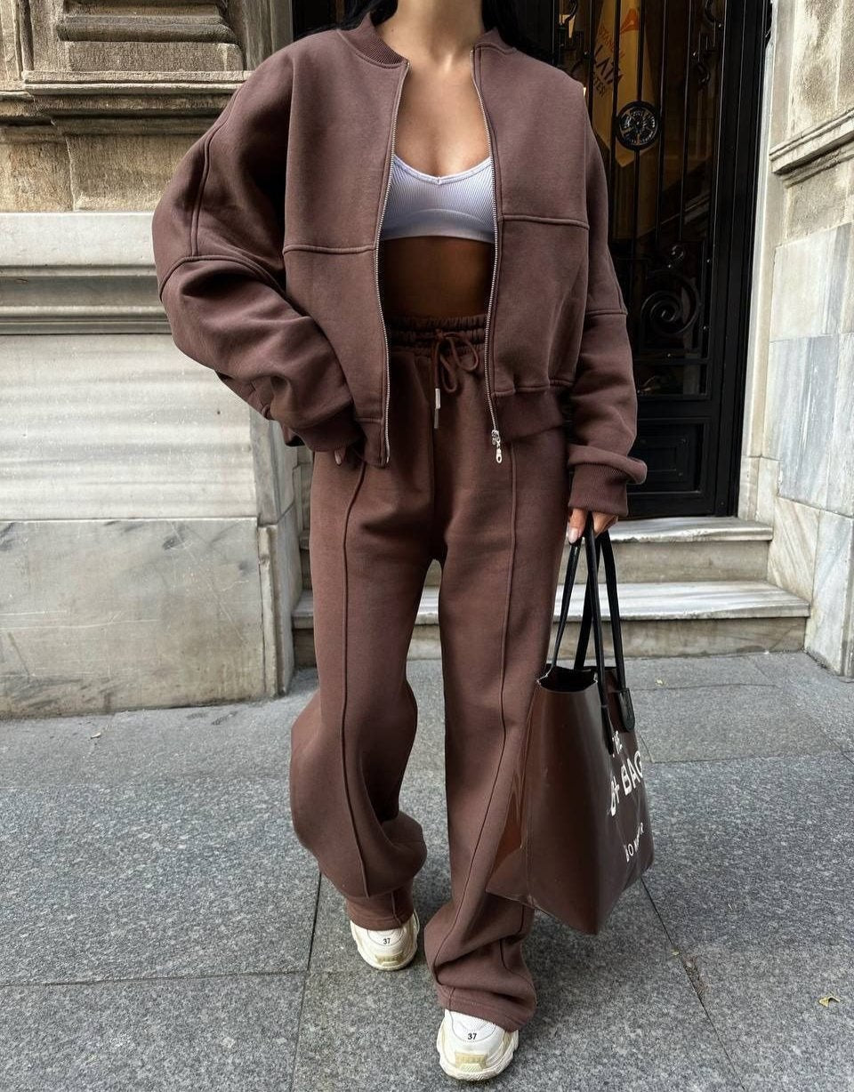 Donna in tuta marrone con top bianco, borsa nera, scarpe da ginnastica bianche, davanti a un edificio storico. Moda casual autunnale.