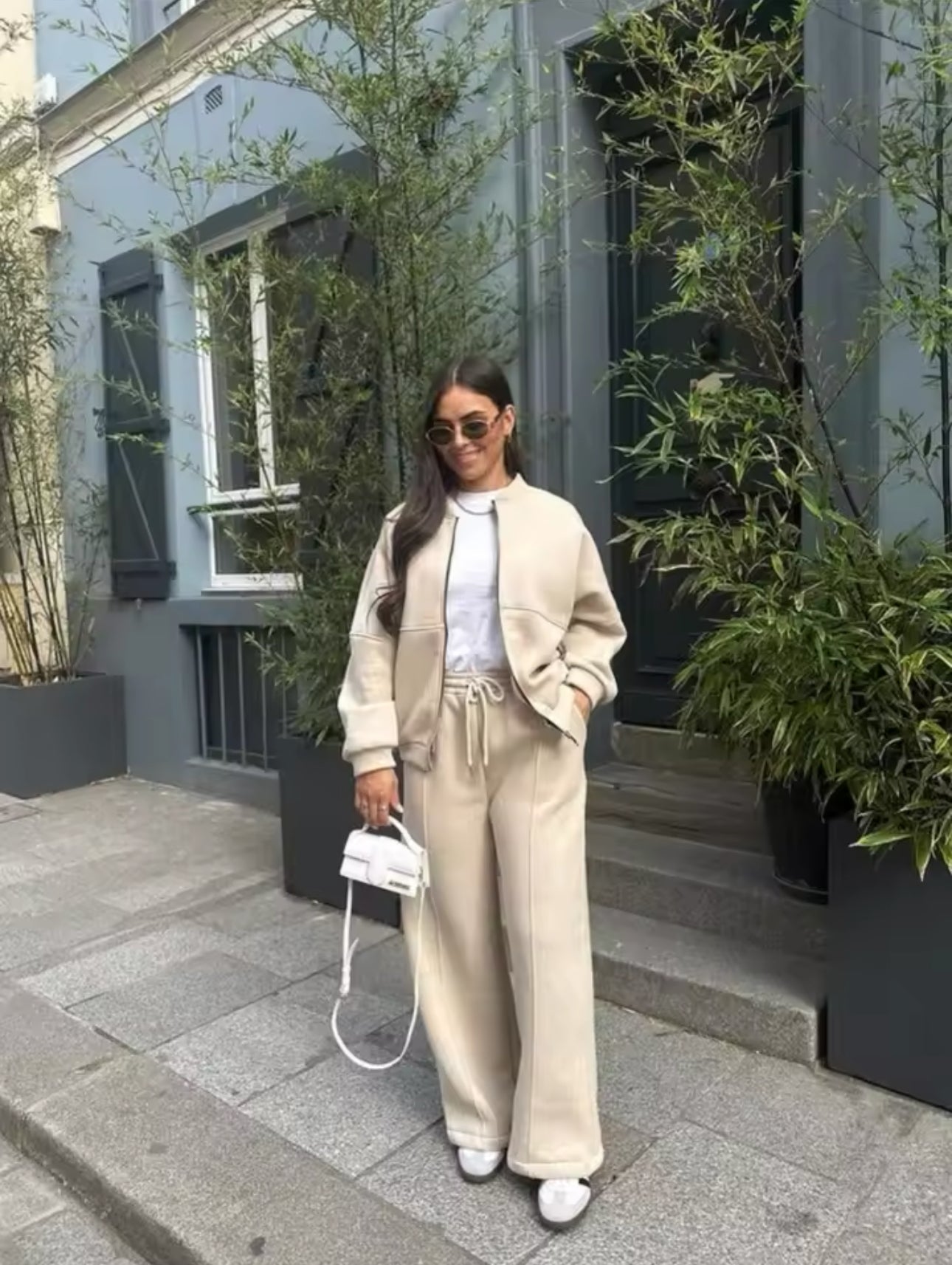 Donna con occhiali da sole indossa completo beige elegante, borsa bianca e scarpe da ginnastica, posa davanti a edificio blu con piante verdi.