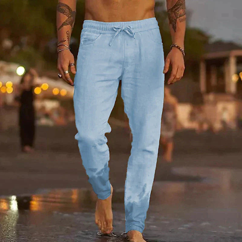 Uomo in pantaloni jogger azzurri cammina sulla spiaggia al tramonto, sfondo sfocato con luci e vegetazione. Moda casual estiva, relax.
