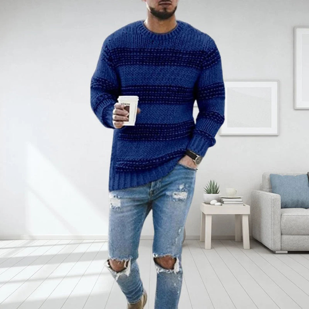 Uomo con maglione blu a righe, jeans strappati, tiene una tazza di caffè. Interno moderno, divano grigio, tavolino bianco. Moda casual maschile.