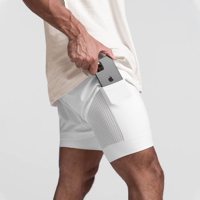Uomo in pantaloncini sportivi bianchi con tasca laterale, inserisce uno smartphone. Abbigliamento fitness, moda sportiva, tecnologia portatile.