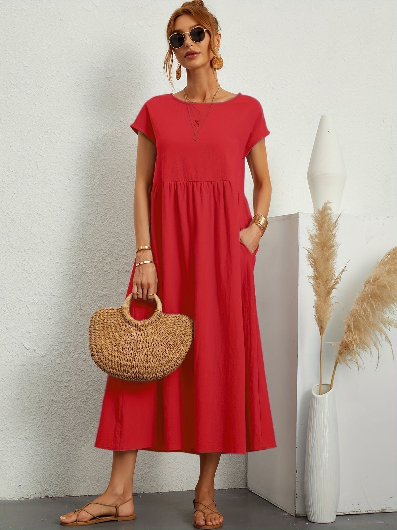 Donna con abito rosso estivo, borsa di paglia e sandali, posa accanto a decorazioni minimaliste. Moda estiva, stile casual chic.