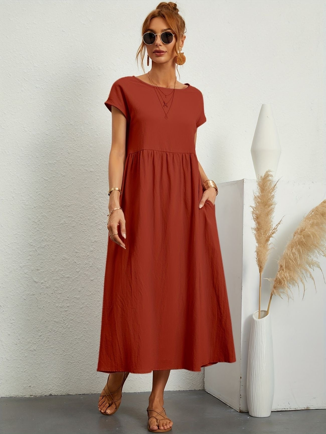 Donna con abito lungo rosso, occhiali da sole e sandali, accanto a vaso bianco con pampas. Moda estiva elegante, stile casual chic.