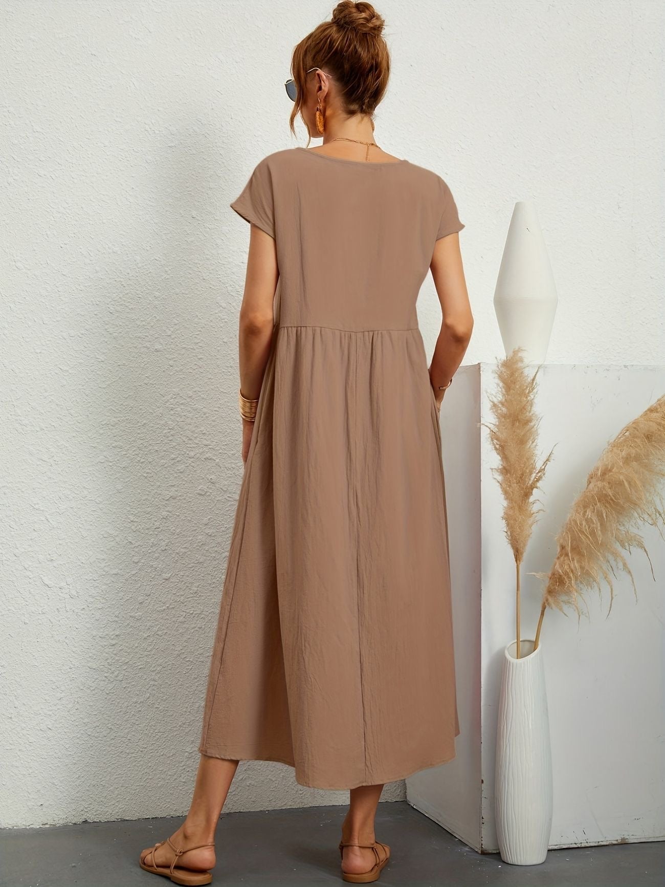 Donna di spalle con abito lungo marrone, sandali beige, accanto a vaso bianco con pampas. Moda estiva, abbigliamento casual elegante.