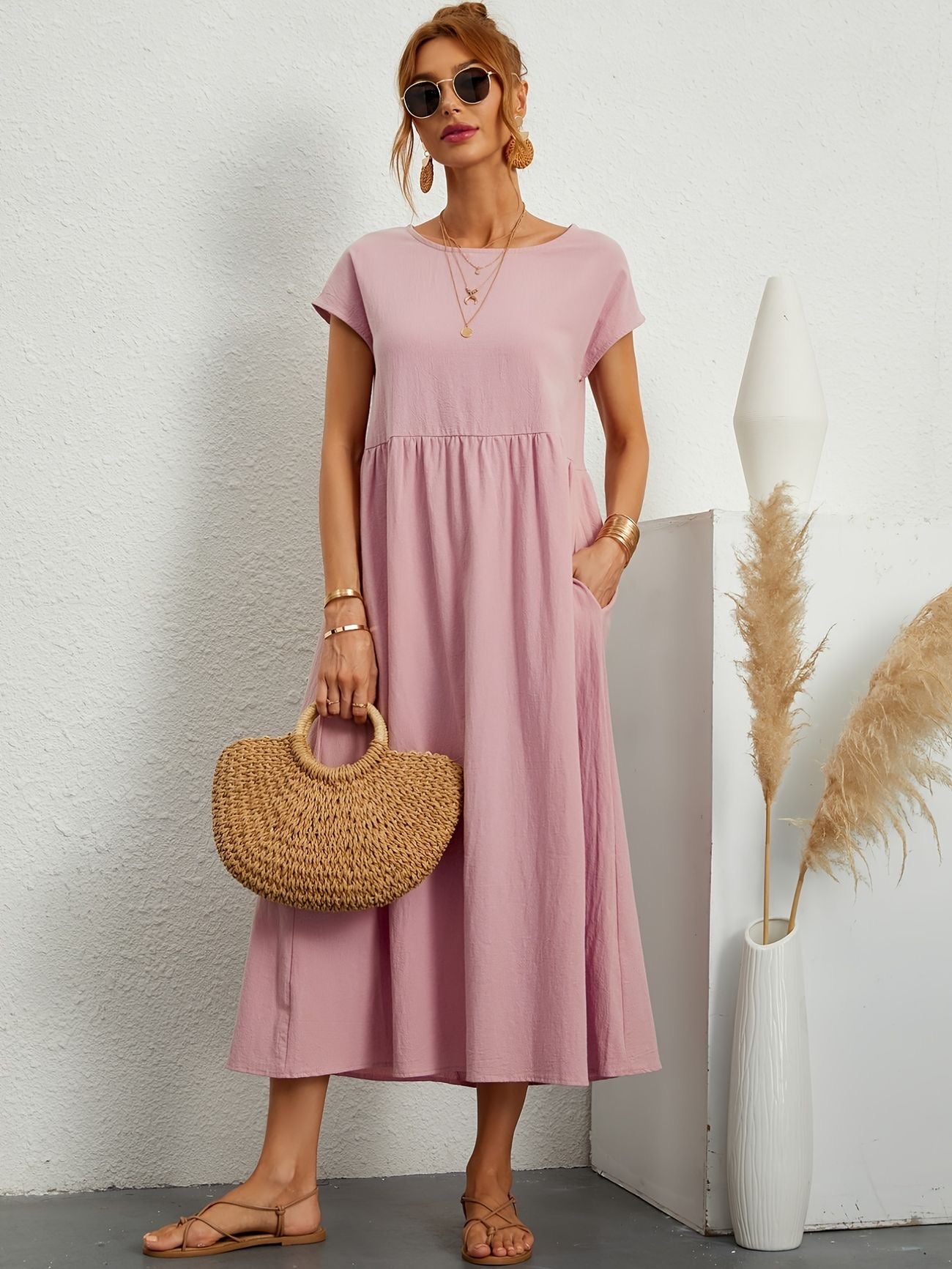 Donna con abito rosa lungo, borsa di paglia, occhiali da sole, sandali beige, accanto a vaso bianco con pampas. Moda estiva elegante e casual.