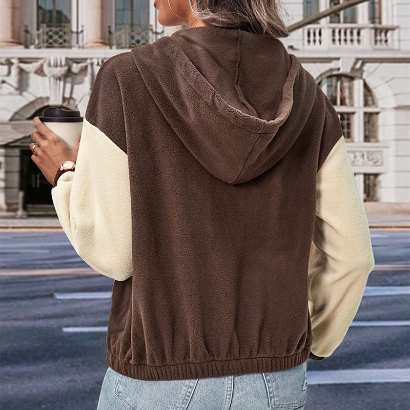 Donna di spalle con felpa marrone e bianca, cappuccio, jeans, tiene una tazza da caffè. Sfondo urbano con edificio storico. Moda casual autunnale.
