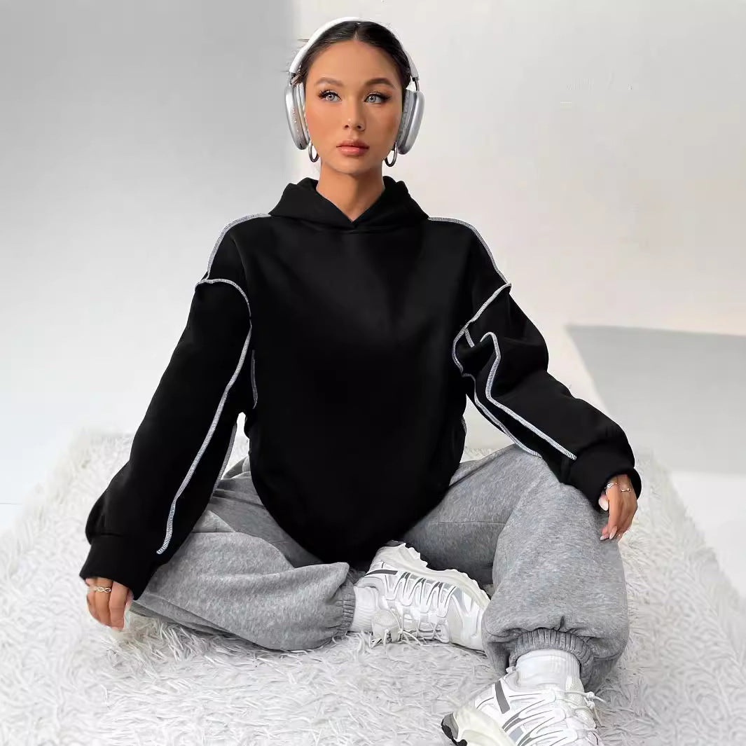 Donna seduta su tappeto bianco, indossa felpa nera, pantaloni grigi, cuffie e sneakers bianche. Moda casual, stile urbano, comfort.