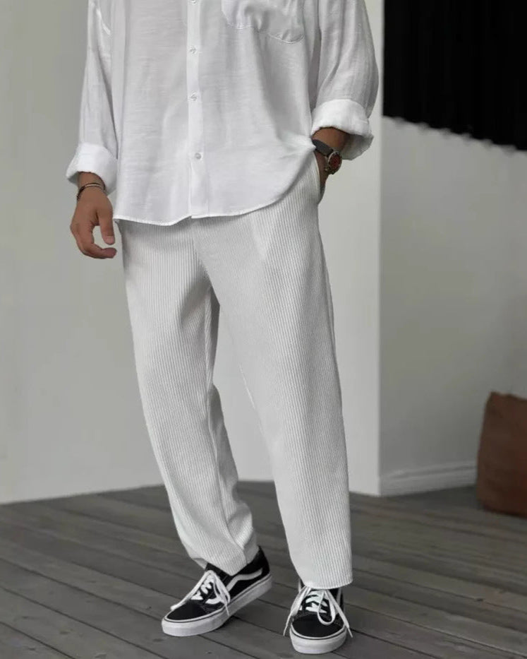 Uomo in camicia bianca e pantaloni bianchi a coste, indossa scarpe da ginnastica nere su pavimento in legno. Moda casual elegante maschile.