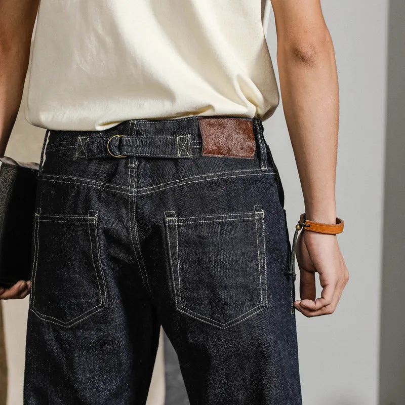 Jeans uomo in denim blu scuro con cuciture a contrasto, cintura in pelle marrone, tasche posteriori, indossati con maglietta bianca. Moda casual maschile.