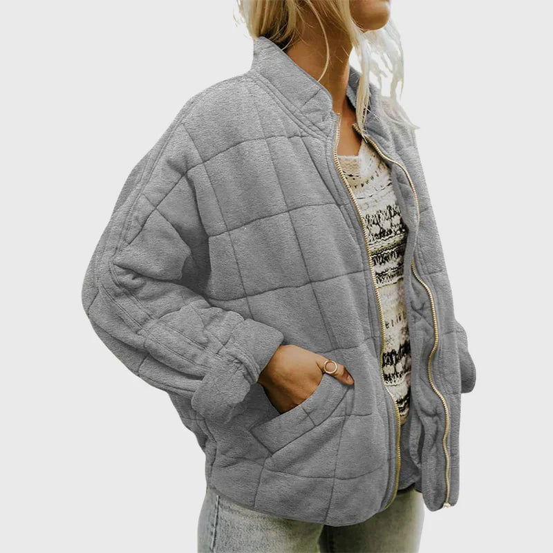 Donna con giacca trapuntata grigia, zip aperta, indossa maglia a fantasia e jeans. Moda autunnale, abbigliamento casual, stile confortevole.