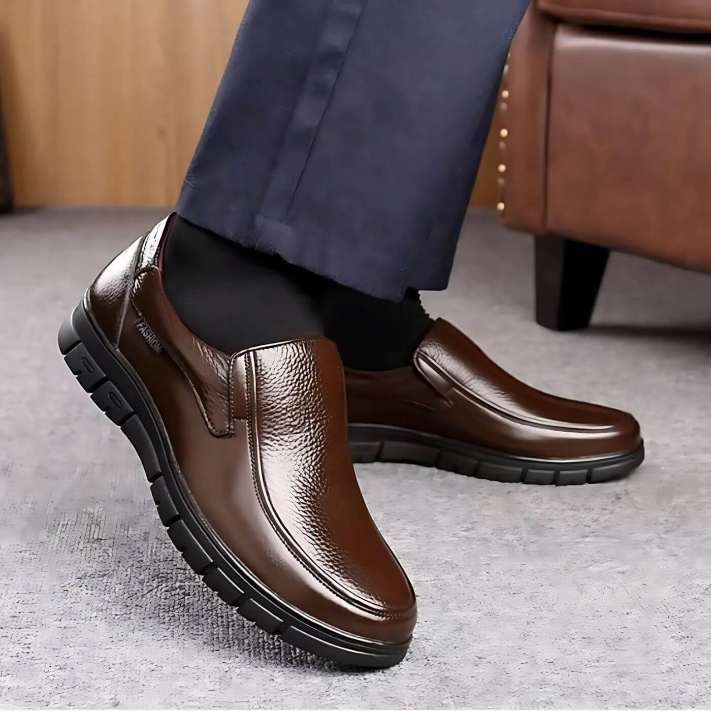 Scarpe eleganti da uomo in pelle marrone, design slip-on, suola nera antiscivolo, indossate con pantaloni blu scuro su sfondo interno moderno.