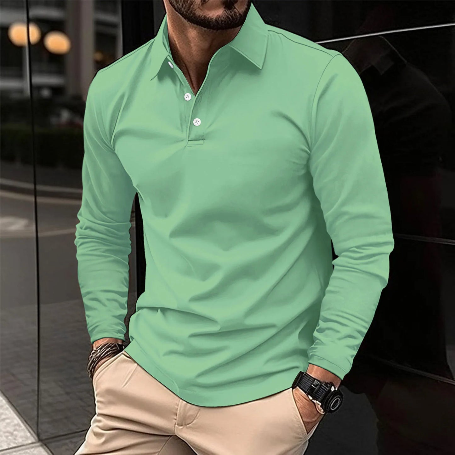Uomo con camicia verde a maniche lunghe, pantaloni beige, orologio nero, in piedi su marciapiede urbano. Moda maschile casual elegante.