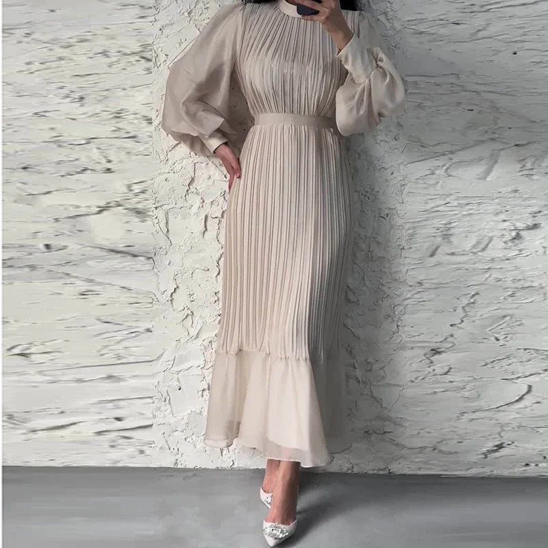 Abito lungo plissettato beige elegante, maniche lunghe, indossato da una donna su sfondo di muro bianco. Stile sofisticato e moderno.