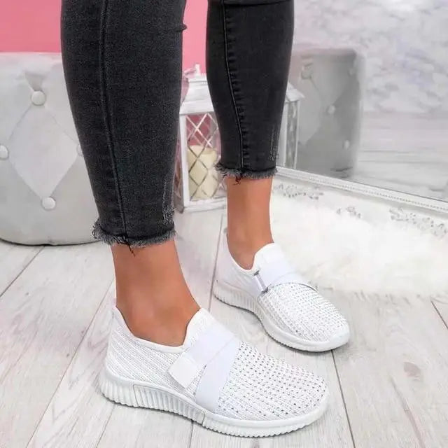 Scarpe da ginnastica bianche da donna, design traforato, indossate con jeans neri. Calzature casual su pavimento in legno chiaro.