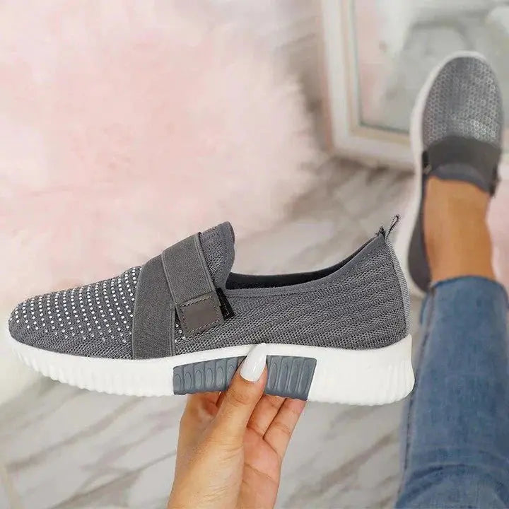 Scarpa sportiva grigia in maglia traspirante con suola bianca, design slip-on, tenuta in mano su sfondo di pavimento marmo e tappeto rosa.