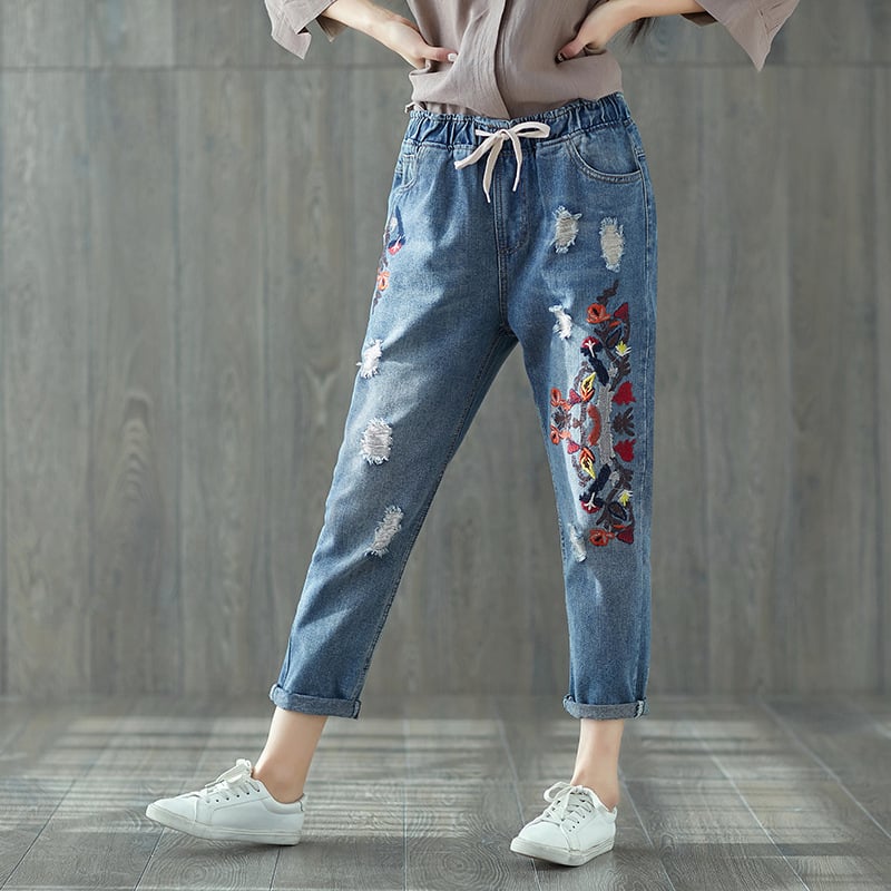 Jeans donna ricamati, strappati, con motivi floreali colorati, indossati con sneakers bianche su pavimento in legno. Moda casual femminile.