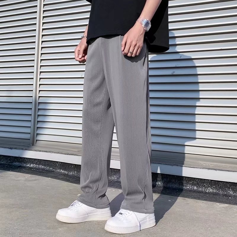 Uomo in pantaloni grigi a pieghe, maglietta nera e scarpe da ginnastica bianche, in piedi davanti a una parete con persiane metalliche.