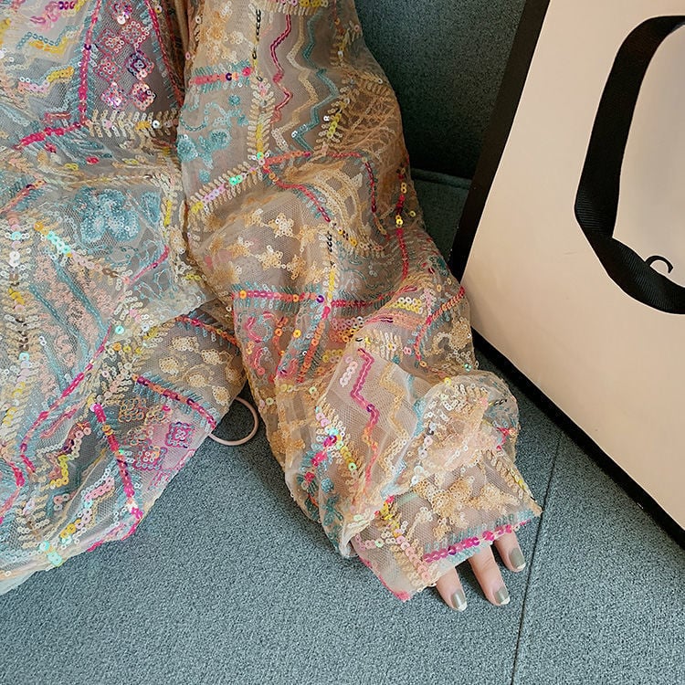Abito in tulle ricamato con paillettes colorate su sfondo beige, dettagli geometrici e floreali, accanto a una borsa bianca su divano blu.