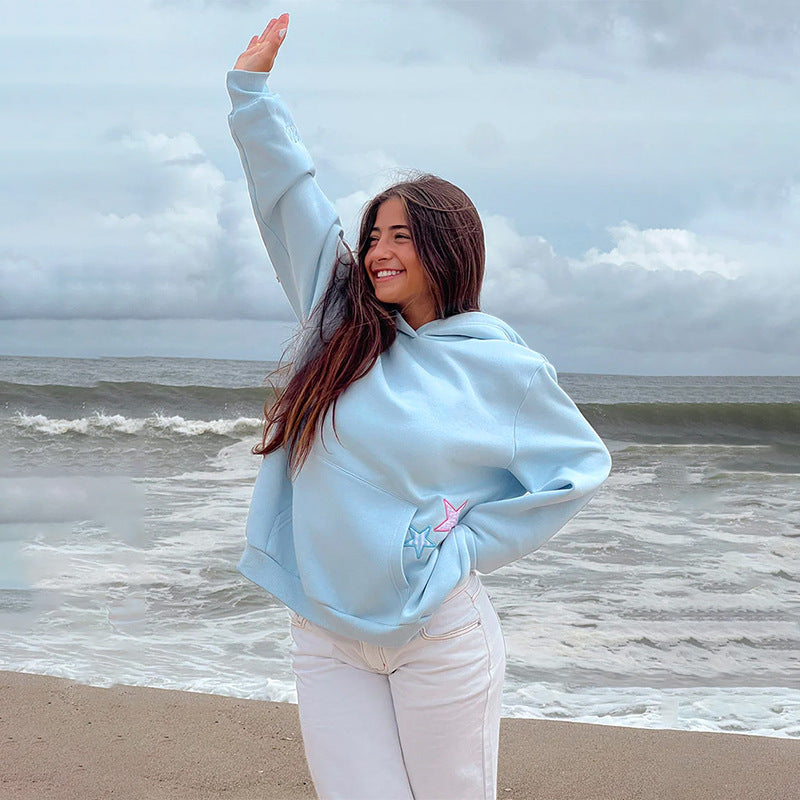 Donna sorridente in felpa azzurra su spiaggia, mare mosso sullo sfondo. Moda casual, abbigliamento comodo, giornata nuvolosa.