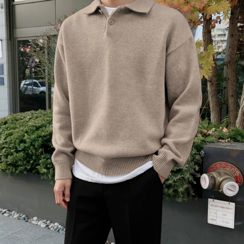 Uomo con maglione beige a collo alto, pantaloni neri, sfondo urbano con piante e foglie autunnali. Moda casual autunno inverno.