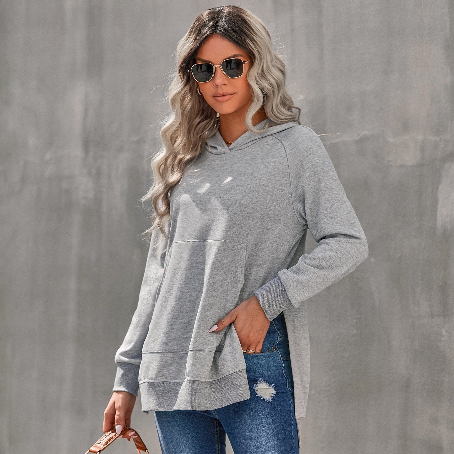 Donna con occhiali da sole indossa felpa grigia con cappuccio e jeans strappati, sfondo grigio. Moda casual, street style, abbigliamento autunnale.