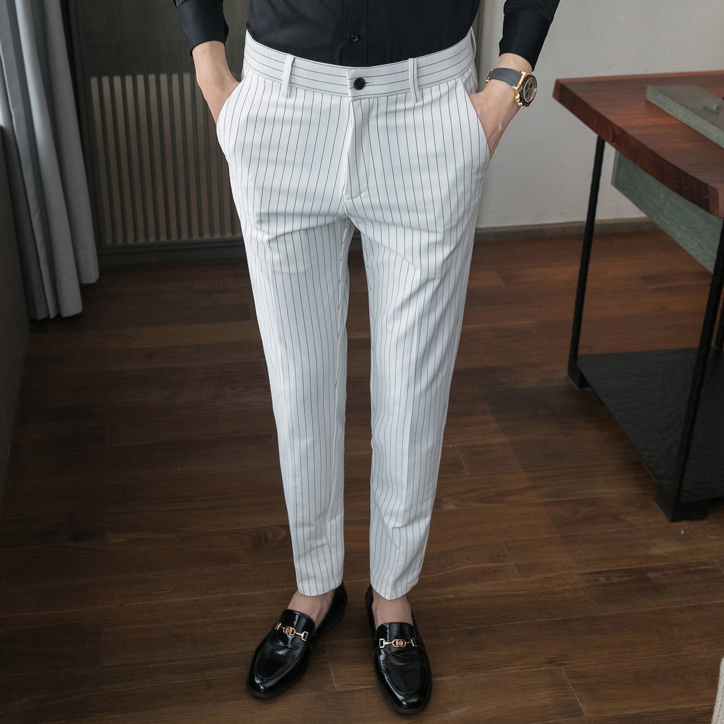 Pantaloni eleganti a righe bianche e nere, indossati con mocassini neri lucidi e camicia nera, su pavimento in legno. Moda uomo formale.