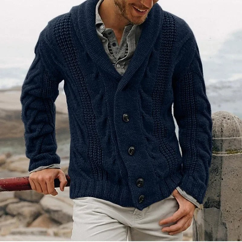 Uomo con cardigan blu scuro a maglia grossa, colletto a scialle e bottoni, indossa camicia grigia e pantaloni chiari, sfondo marino.