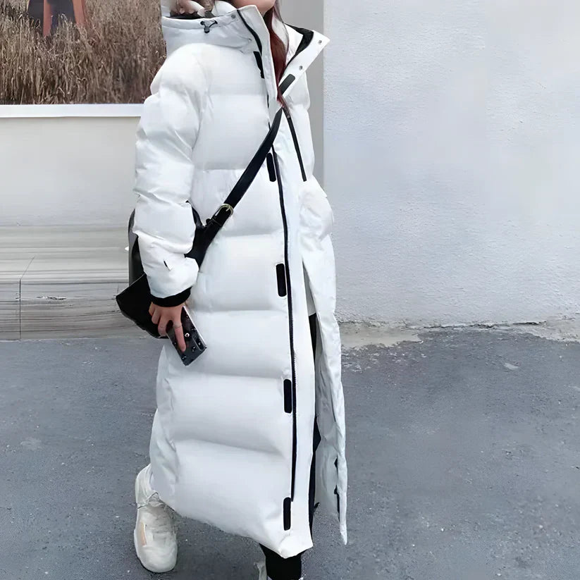 Donna con lungo piumino bianco invernale, cappuccio e dettagli neri, cammina su marciapiede grigio. Stile moda inverno, abbigliamento caldo.