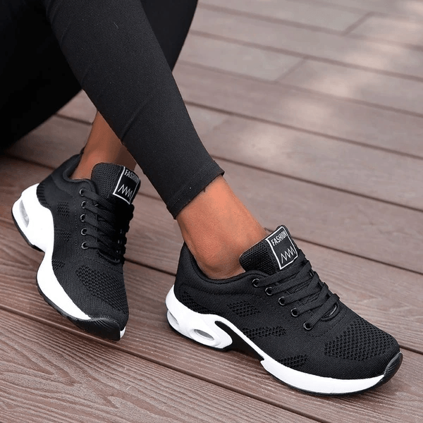 Scarpe da ginnastica nere da donna su pavimento in legno, design moderno e traspirante, ideali per sport e tempo libero.