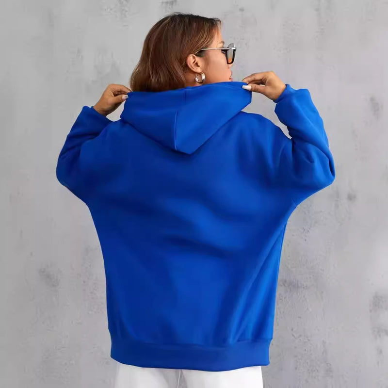 Donna con felpa blu oversize, cappuccio alzato, sfondo grigio. Moda casual, abbigliamento autunnale, stile urbano, comfort quotidiano.