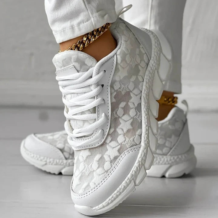 Scarpe da ginnastica bianche con motivo floreale, suola spessa e dettagli dorati, indossate con pantaloni bianchi. Stile casual e moderno.