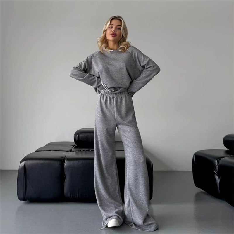 Donna in tuta grigia elegante, posa in salotto moderno con divano nero. Moda casual, abbigliamento comodo, stile autunnale.