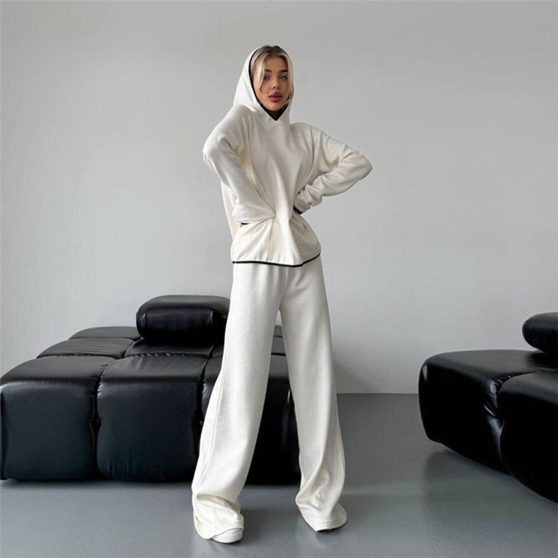 Donna in tuta bianca con cappuccio, posa in salotto moderno con divani neri. Moda casual, abbigliamento comodo, stile minimalista.