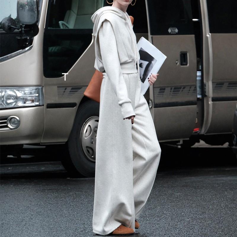 Donna in tuta bianca elegante, cappuccio e pantaloni larghi, tiene una rivista. Sfondo con furgone grigio. Moda urbana, street style.