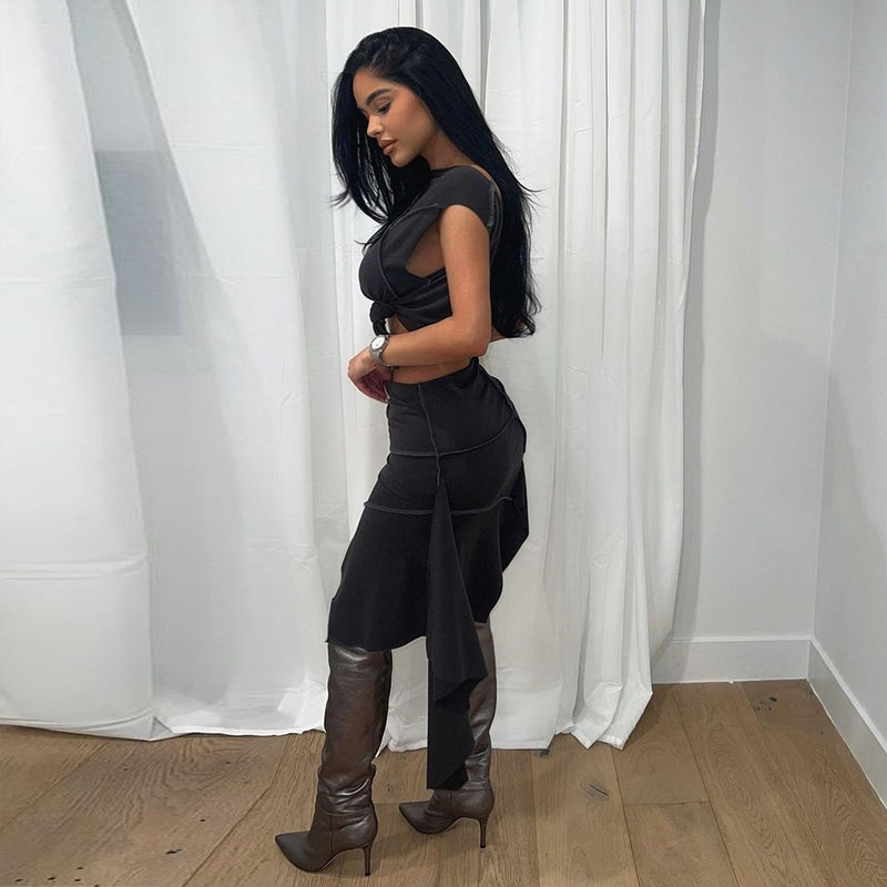 Donna in abito nero elegante e stivali marroni, posa di profilo in una stanza con pavimento in legno e tende bianche. Moda femminile contemporanea.