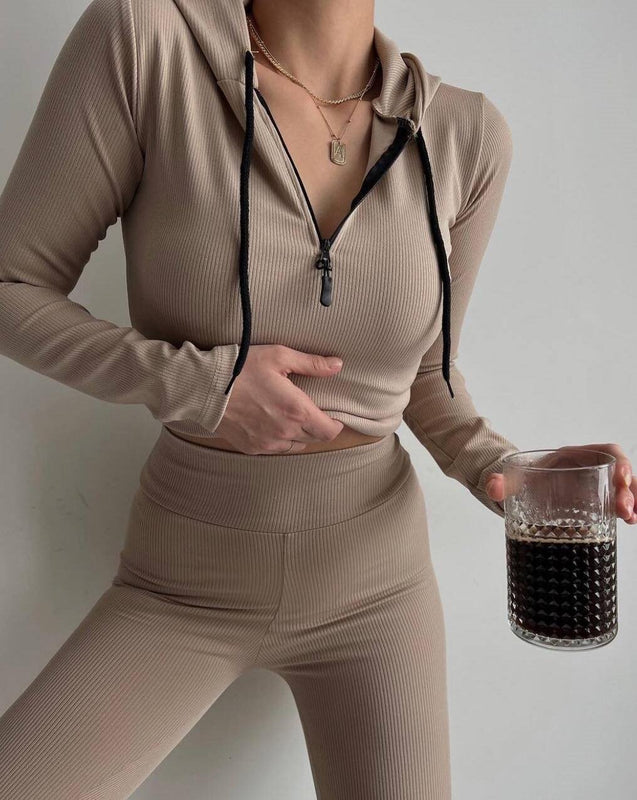 Donna in abbigliamento sportivo beige, felpa con cappuccio e pantaloni a vita alta, tiene un bicchiere di bevanda scura. Moda fitness casual.