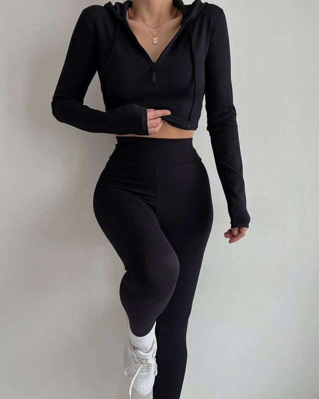 Donna in abbigliamento sportivo nero, con top a maniche lunghe e leggings, posa su sfondo bianco. Moda fitness, abbigliamento activewear elegante.
