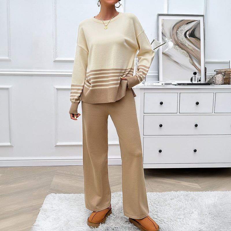 Donna in salotto indossa completo beige a righe, maglione e pantaloni larghi, su tappeto bianco. Arredamento moderno con cassettiera e decorazioni.