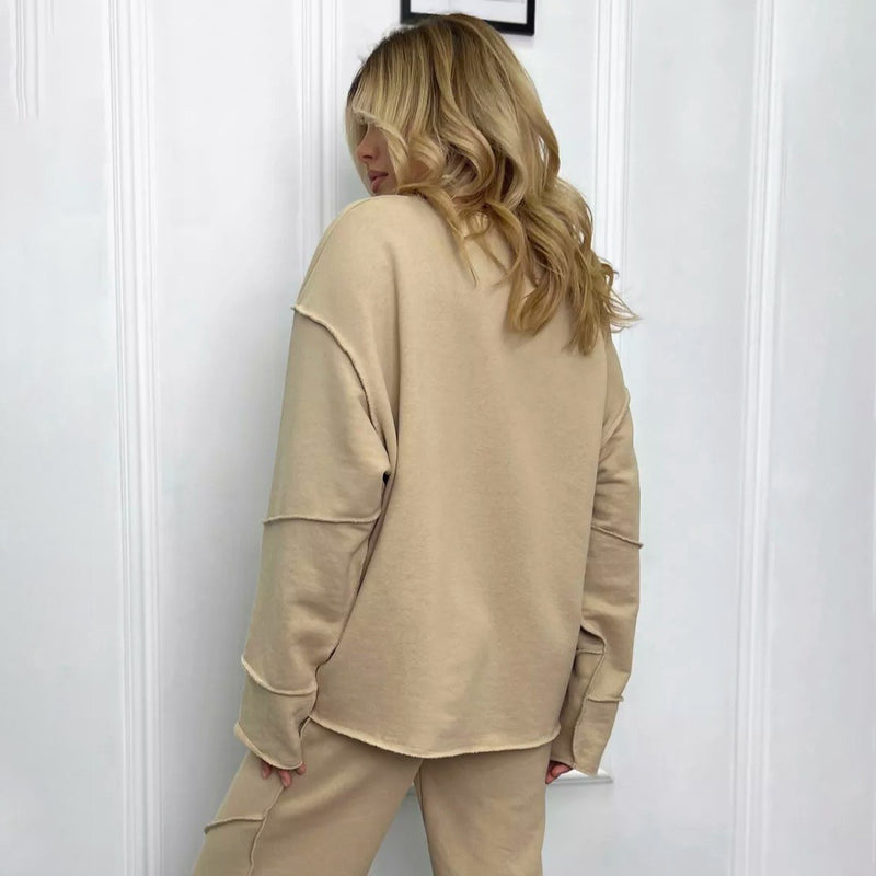 Donna di spalle con capelli biondi indossa tuta beige, composta da felpa e pantaloni, davanti a parete bianca. Moda casual, abbigliamento comodo.