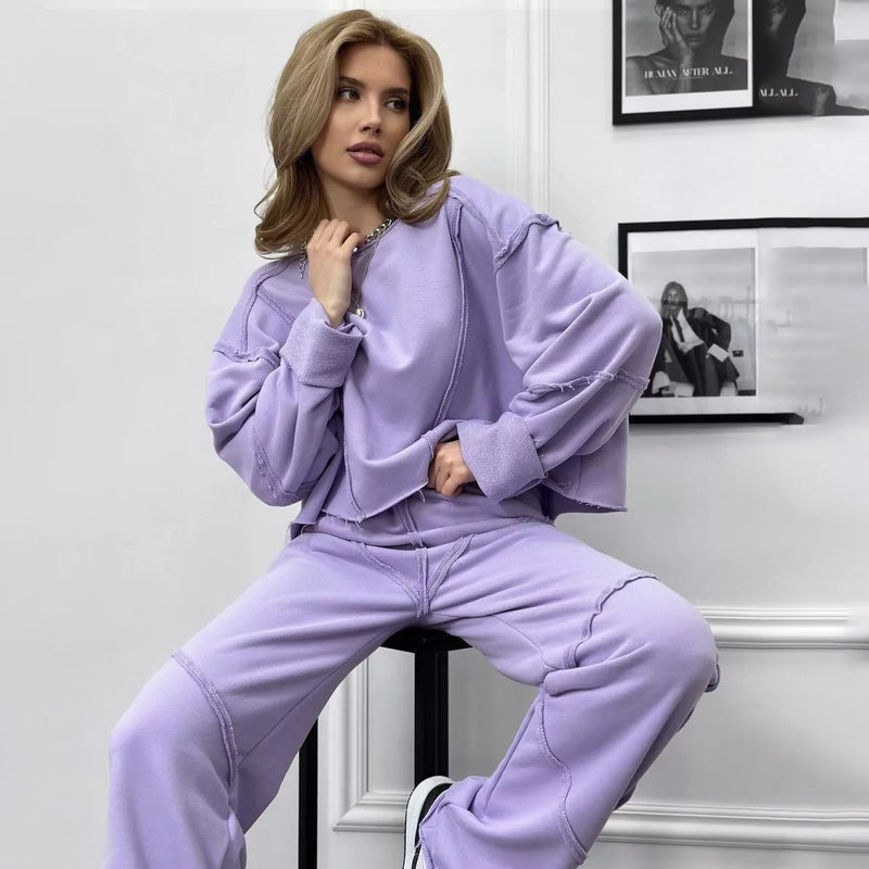 Donna in tuta sportiva lilla, seduta con posa rilassata. Abbigliamento casual, moda femminile, stile urbano. Sfondo bianco con quadri.