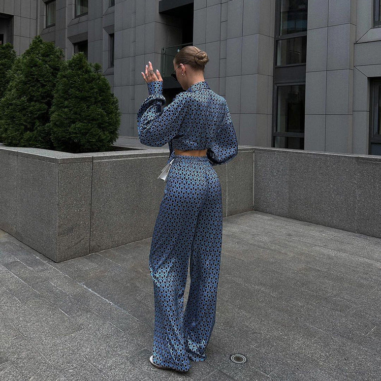 Donna di spalle in tuta elegante blu con motivo geometrico, cammina su marciapiede urbano. Edificio moderno grigio sullo sfondo. Moda urbana chic.