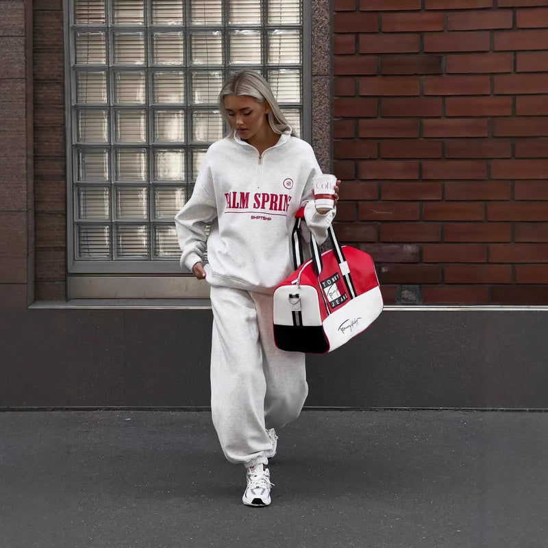 Donna con tuta grigia "Palm Springs", borsa sportiva rossa e bianca, cammina su marciapiede urbano. Stile casual, moda streetwear.