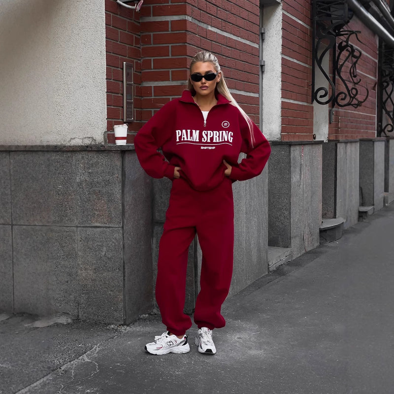 Donna con tuta rossa "Palm Spring", occhiali da sole neri, scarpe da ginnastica bianche, posa su marciapiede urbano. Moda streetwear autunnale.